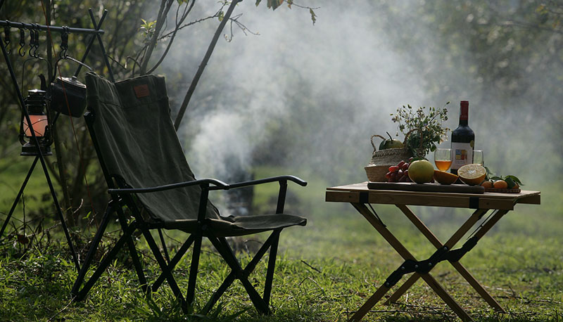 A folding chair, a table with food and drink, barbeque smoke in the background