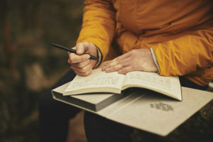 Arms and hands of a person wearing orange robes, holding a pen and notebook, ready to write.