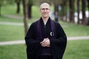 Koshin wearing black monk robes with grass and tree trunks