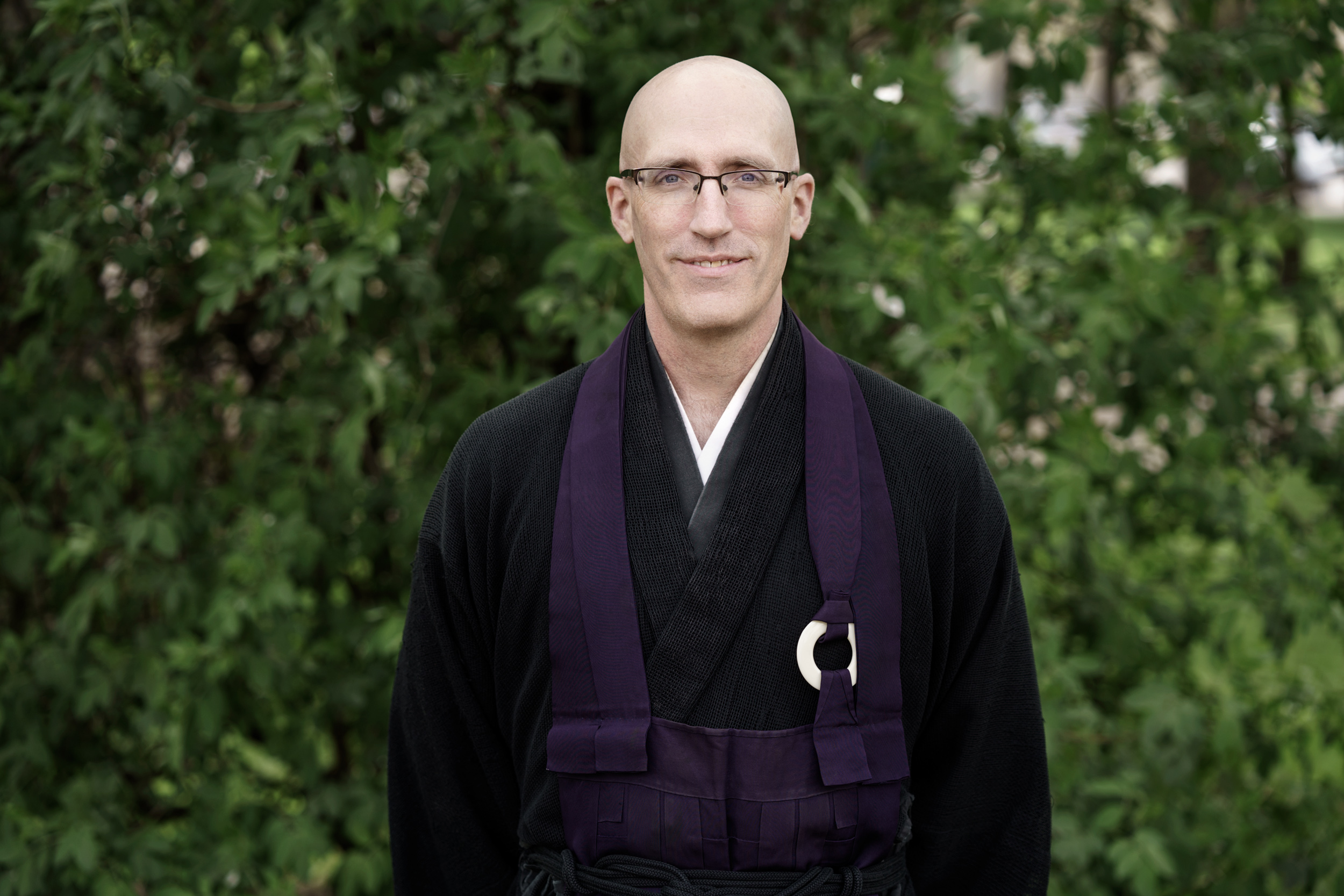 Koshin, wearing his monk's robe, standing in front of green bushes.