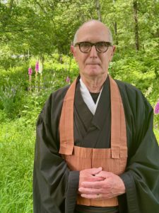 Eko Jeff Kelley in black robes standing outside in front of greenery.