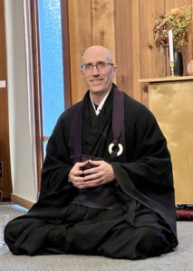 Koshin in black monk robes sitting on the floor in the zendo with his fingers touching.