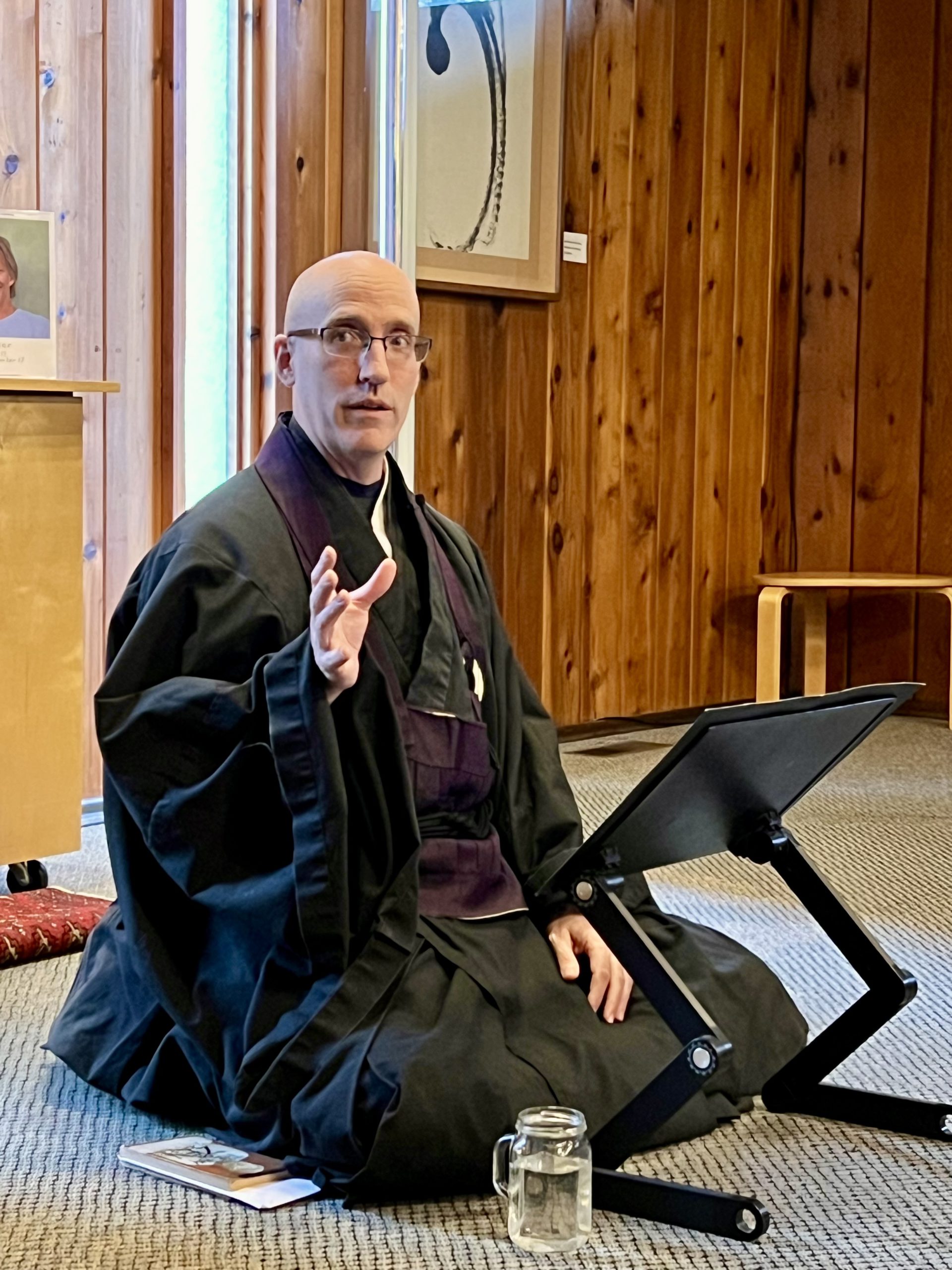 Koshin in a black monk's robe, sitting on the floor, gesturing.